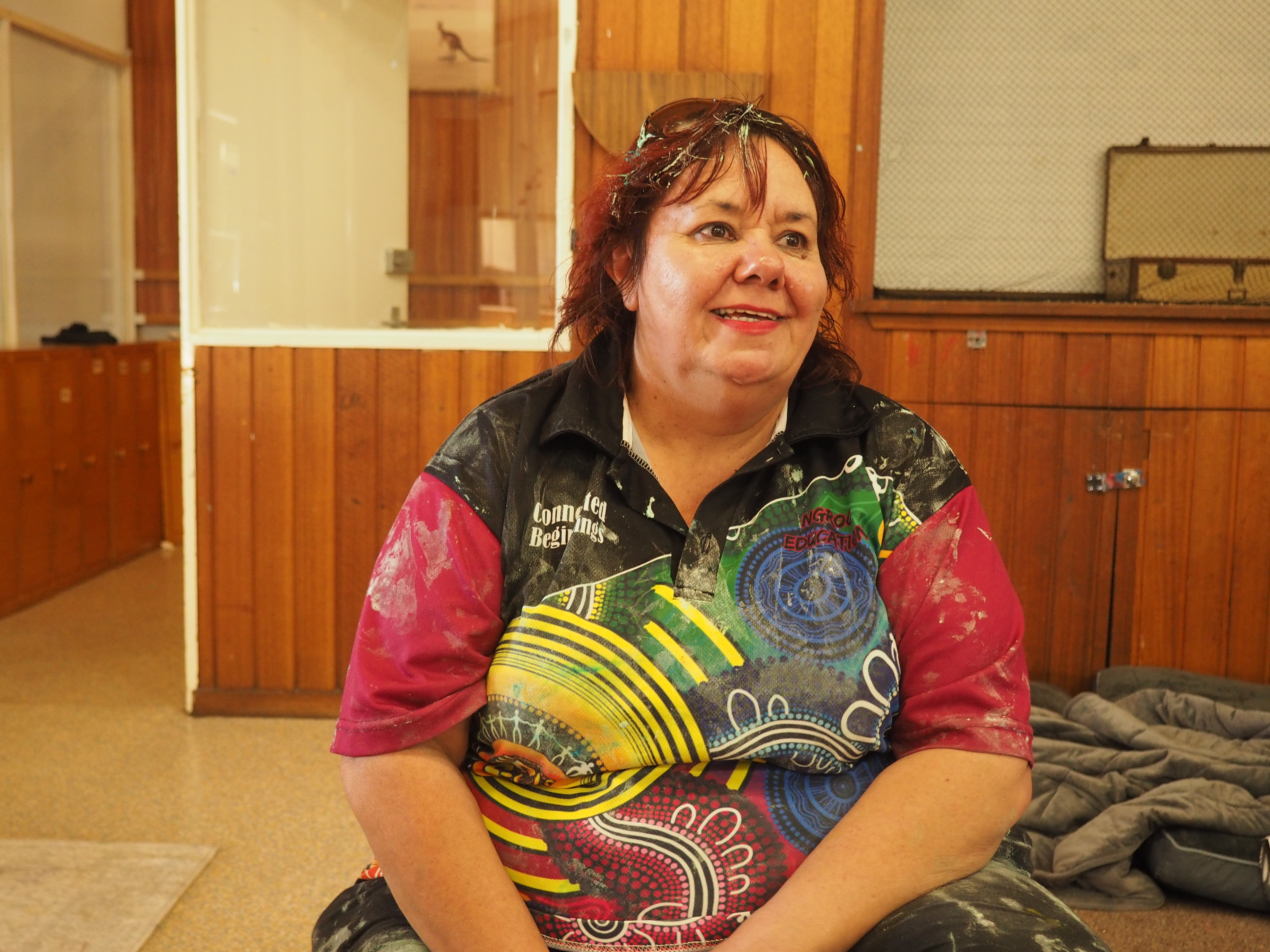 Image of a smiling, woman with red hair sitting cross legged on the floor wearing a brightly coloured Aboriginal inspired short sleeved tshirt with paint stains