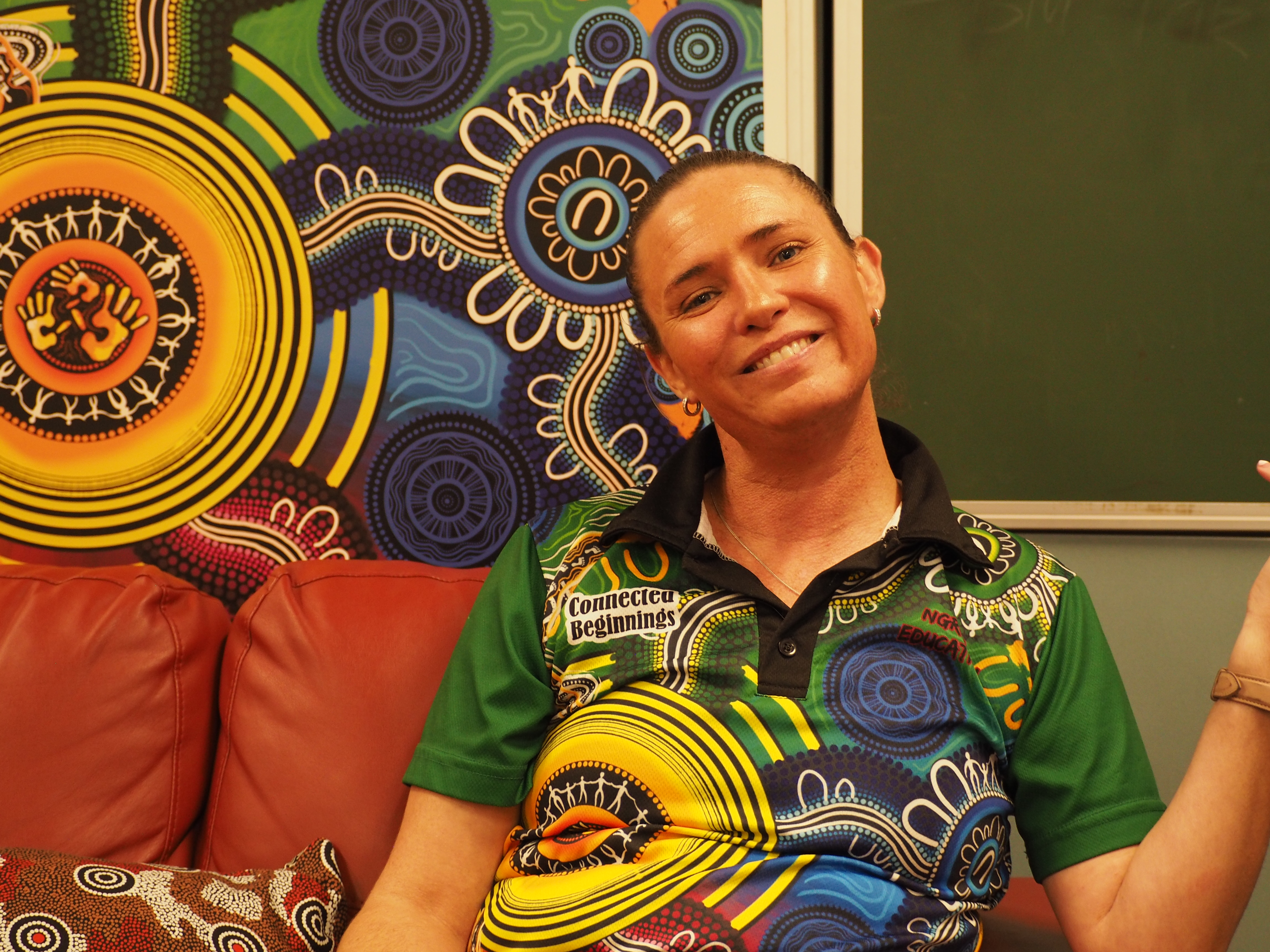 Smiling woman with her dark hair tied up wearing small earrings and wearing a Connected Beginnings Aboriginal colourful t-shirt