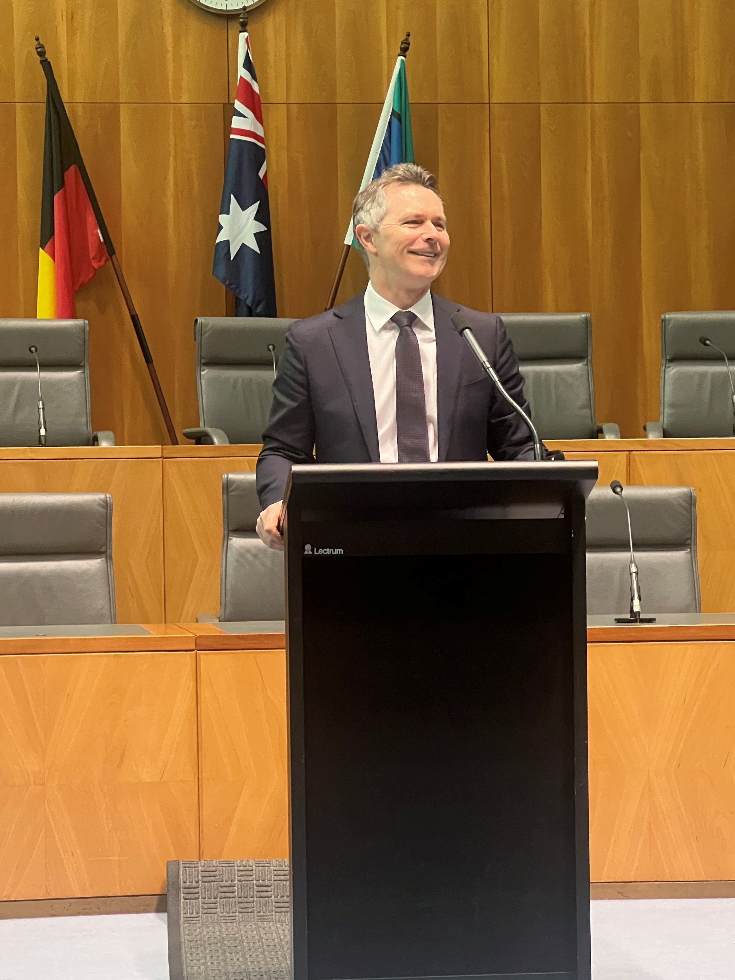 Image of Minister Clare. A man in a business suit, tie and white collared shirt with short grey hair standing behind a podium giving a speech.