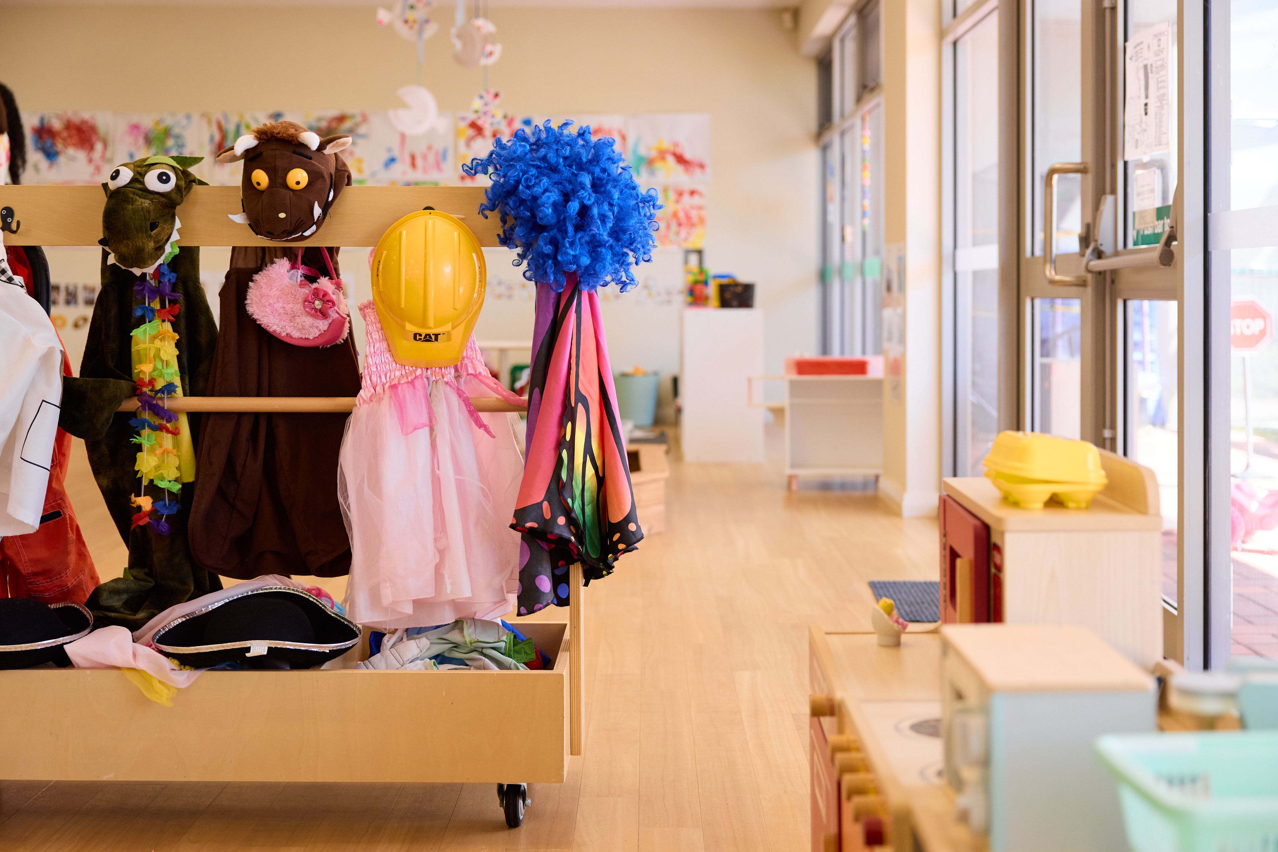 A preschool classroom with dress up clothes, hats and wigs hanging up and a small toy kitchen.