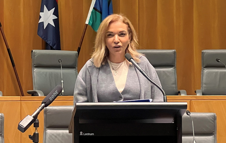 Image of Minister Aly. A woman with shoulder length blonde hair wearing a white top and grey jacket standing behind a podium giving a speech.