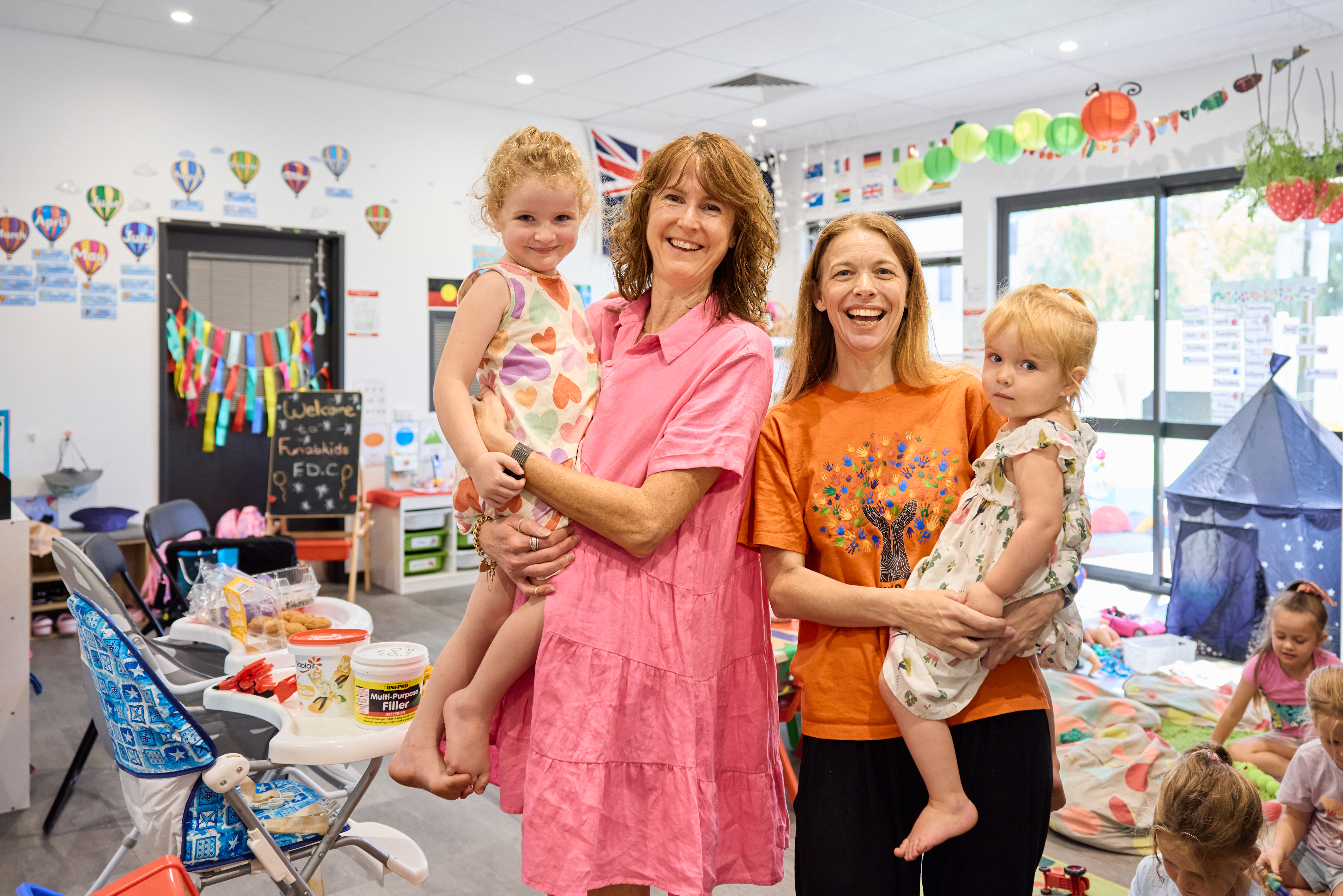 DFC WA early childcare centre classroom with two teachers smiling, holding two young children.