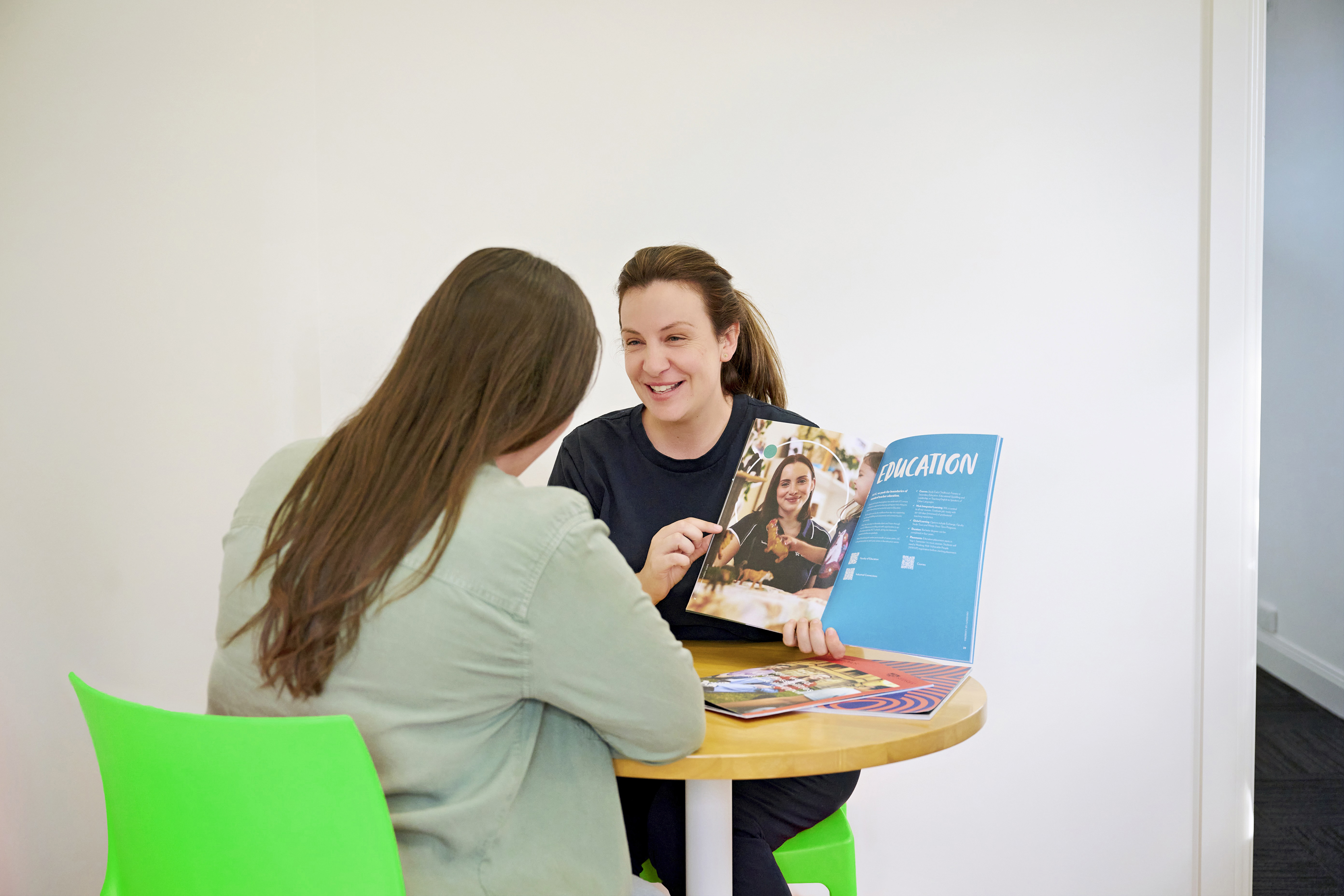 Two women talking and sitting at a desk holding a book.