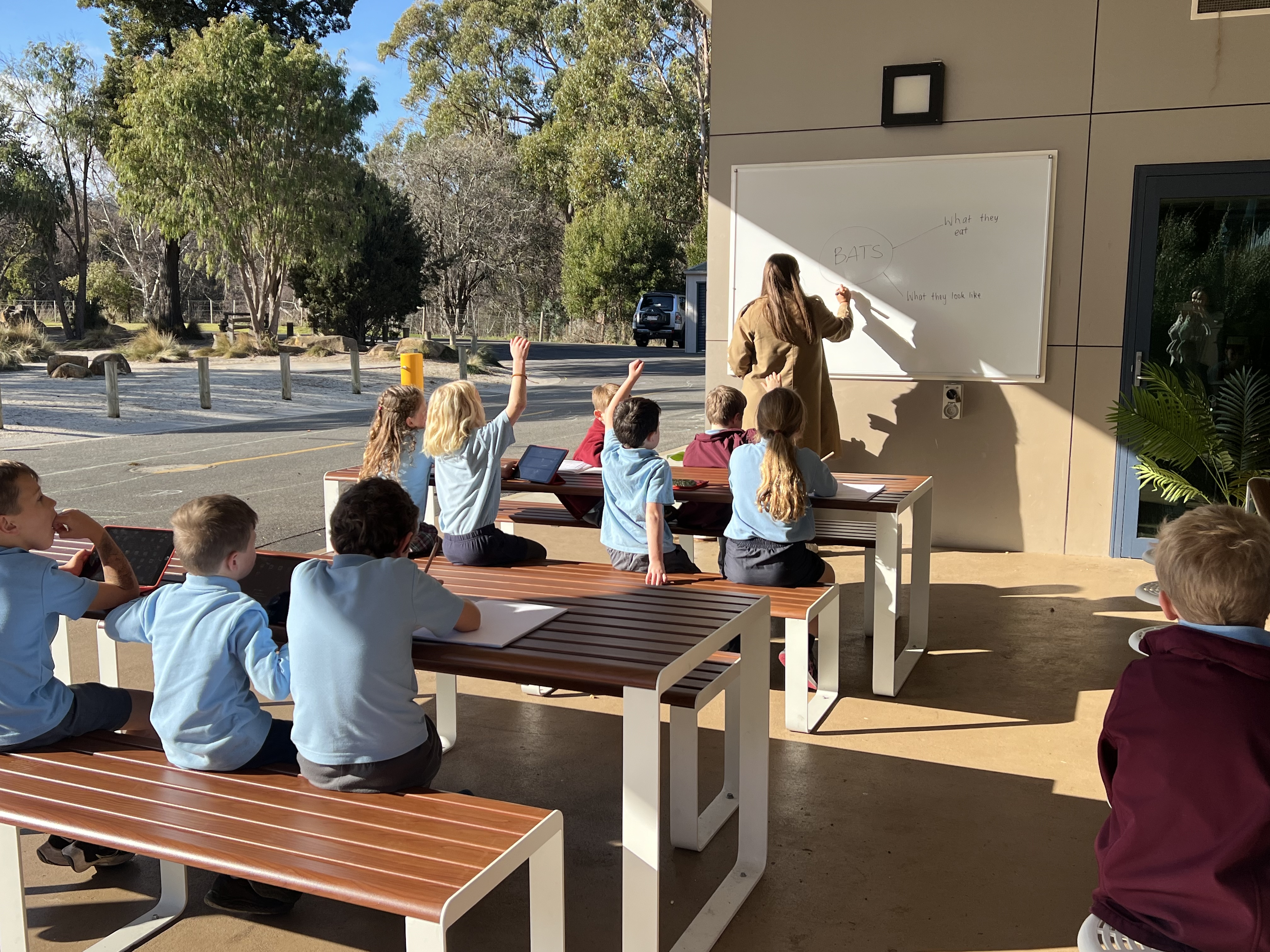 kids sitting at their desks outside looking at the teacher writing on a white board