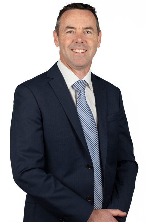 Image of Dr Peter Nolan from the waist up, slightly turned to the left smiling at the camera. Dr Nolan is in a blue suit with a white shirt and blue and white tie
