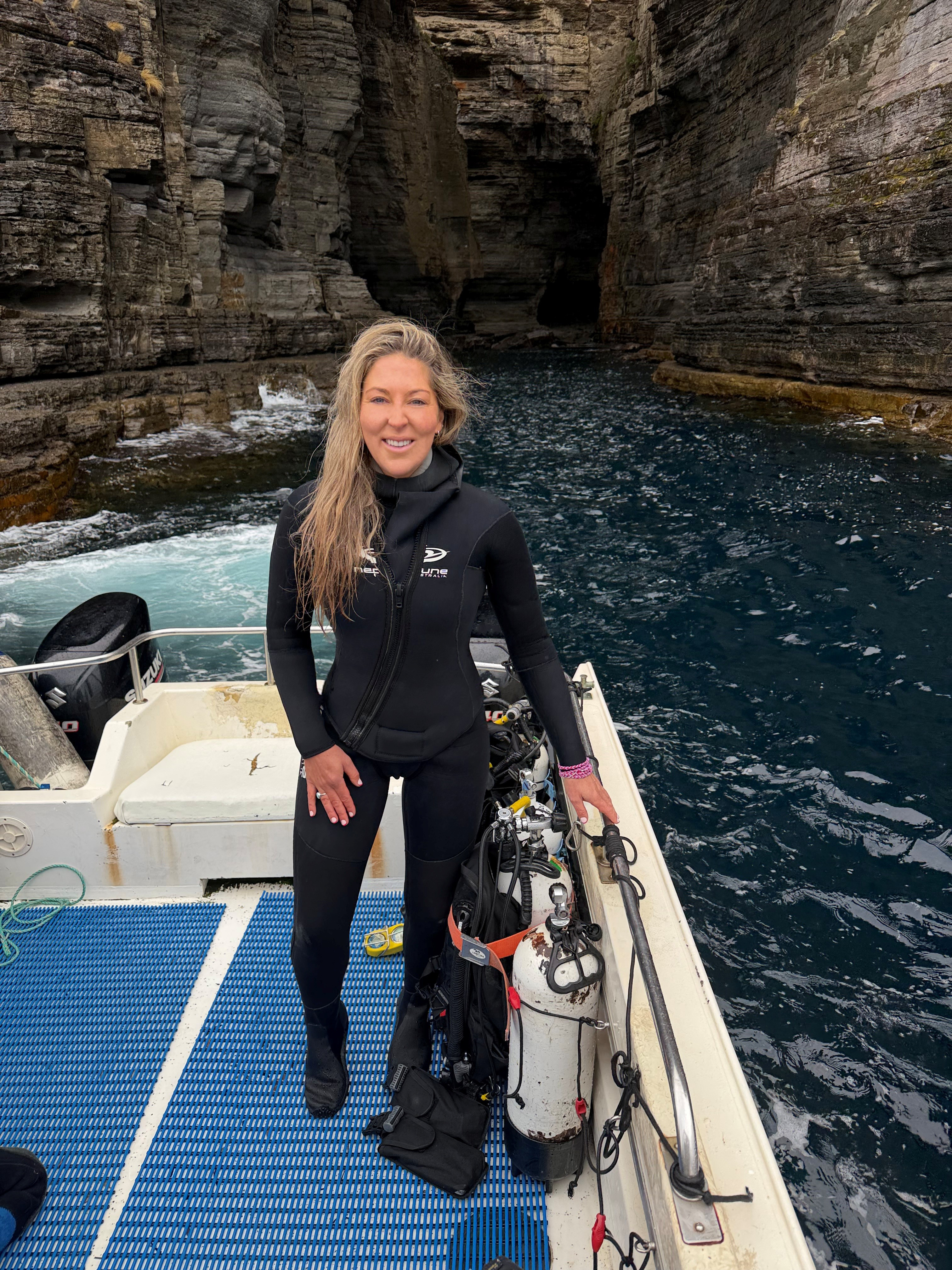 Antonia Cooper smiling dressed in a full wetsuit on a boat floating in the ocean.