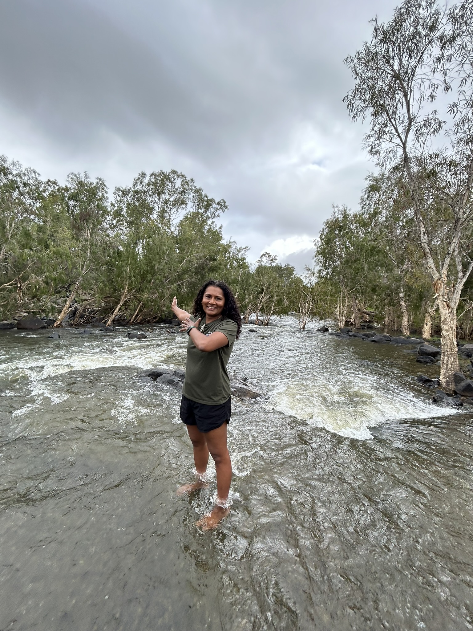 Jo Owens standing in a river stream smiling with trees on the edge of each side of the river