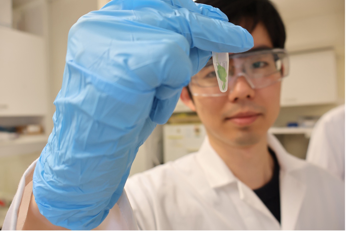 PhD candidate Shunichiro Tomura wearing glasses, a white coat holding a test tube.