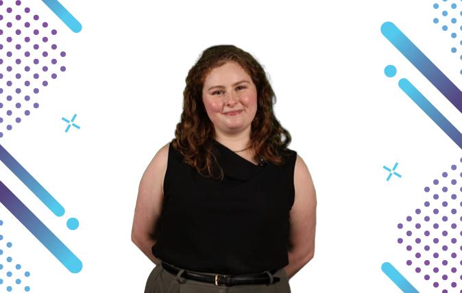 Photo of Gaby with long wavy hair, clasping her hands behind her back, wearing a black, sleeveless top.  She is standing in front of a backgrounds with spots and stripes.