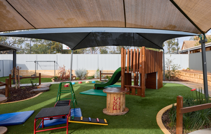 Image of playground with shade sails and play equiptment on grass with high fences in the background